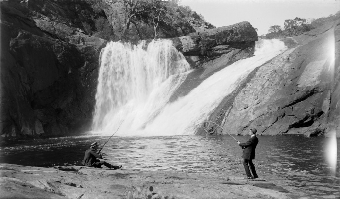 Black and white photo of a man fishing in the pool under a waterfall