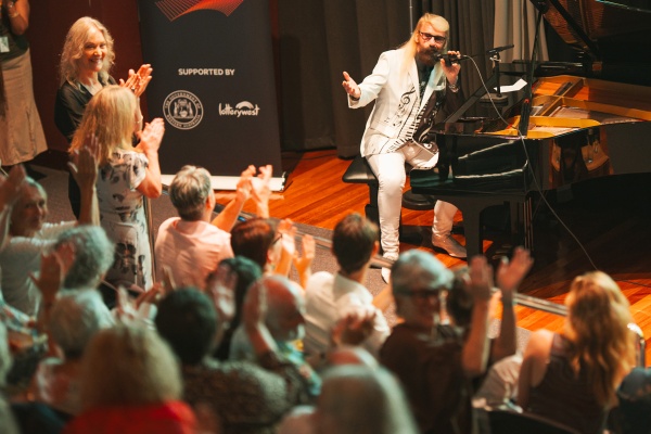 man sitting at piano and speaking into microphone addressing clapping crowd in a theatre