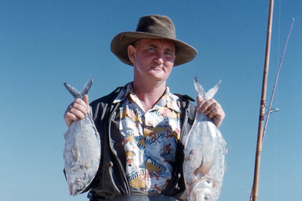 A man in a hat and shorts stands on white sand holding two silver fish by their tails