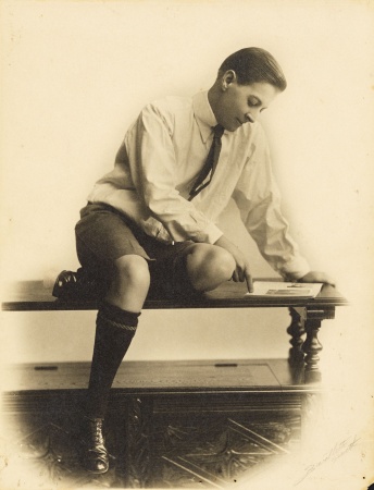 Studio portrait of Effie Fellows seated on a dumb waiter wearing shorts and a shirt and tie