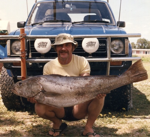 A man crouches in front of a 4wd holding a large fish almost as long as the car is wide