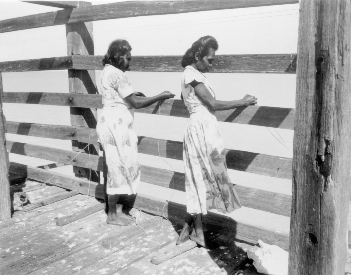 Two Aboriginal women fish drop fishing lines through wooden slats off what looks like a jetty