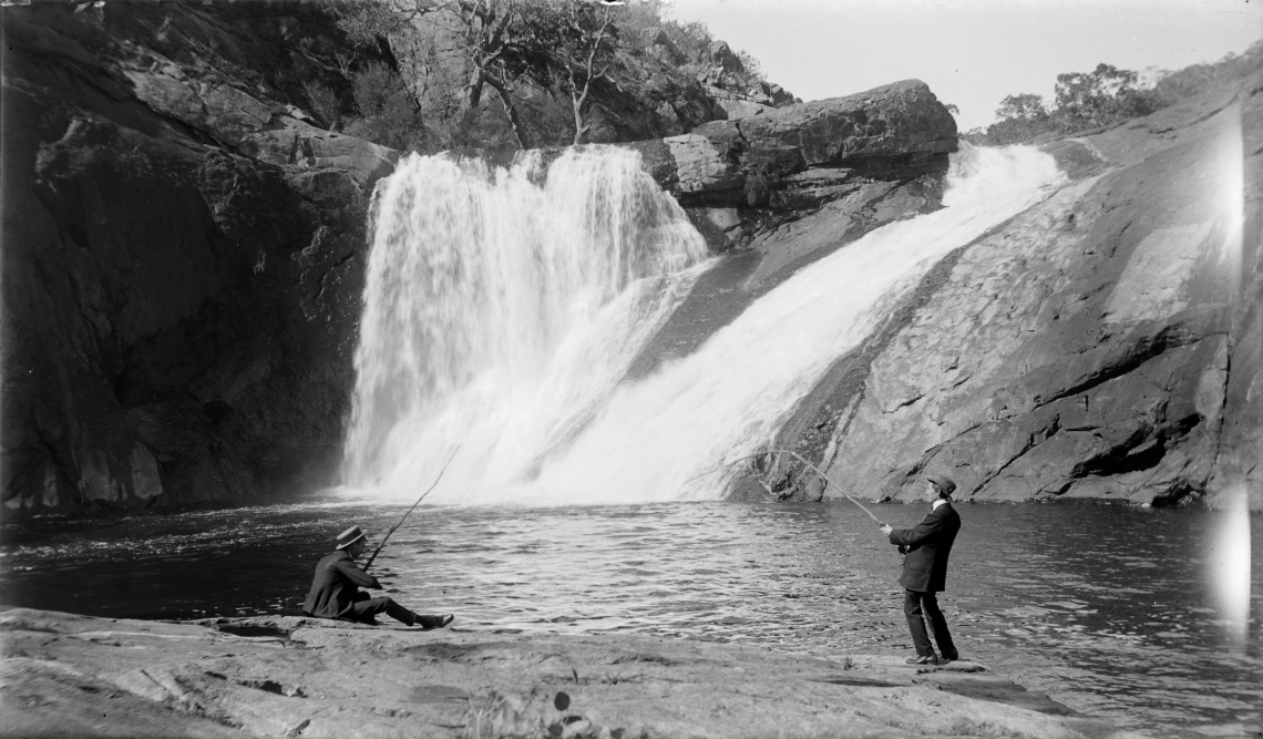 Black and white photo of a man fishing in the pool under a waterfall