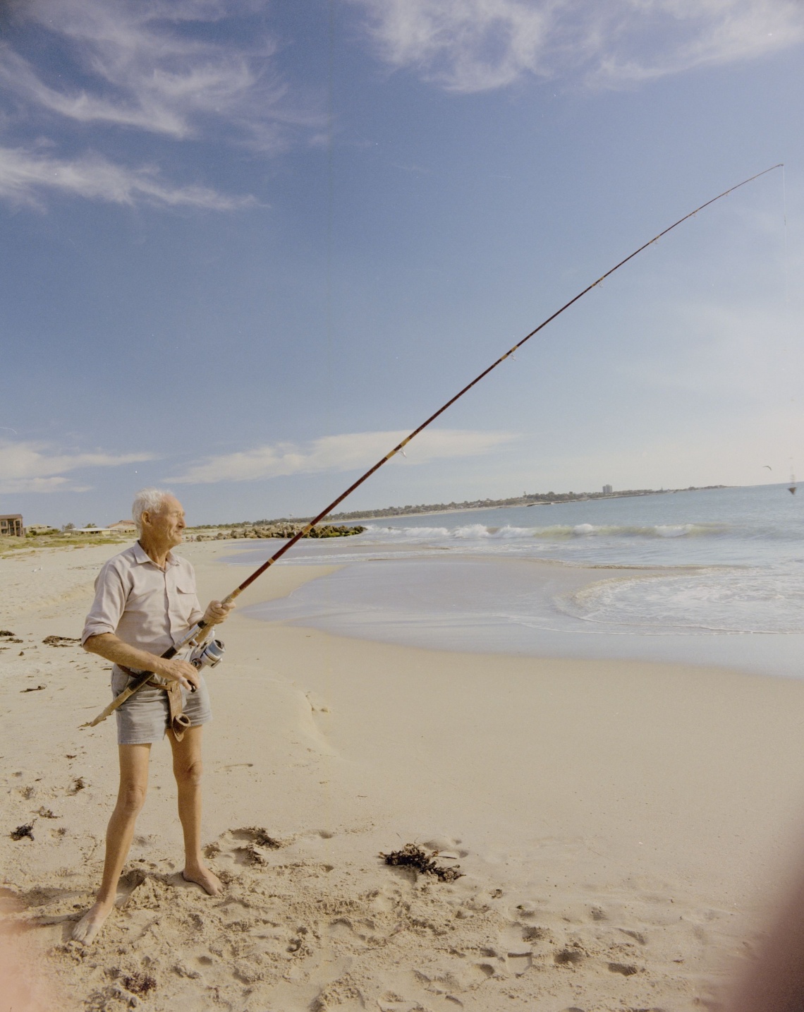 A man in shorts and a button down shirt fishes from the beach