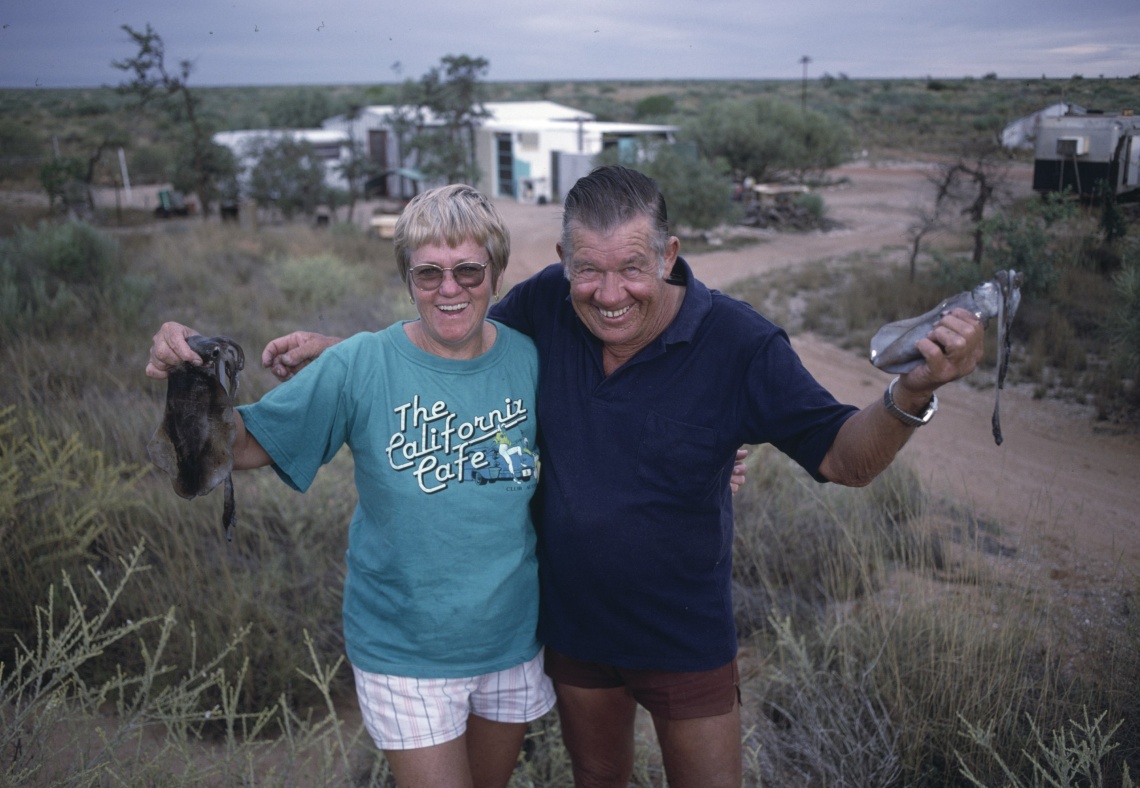 An older couple have their arms around each other each holding a squid in their free hand