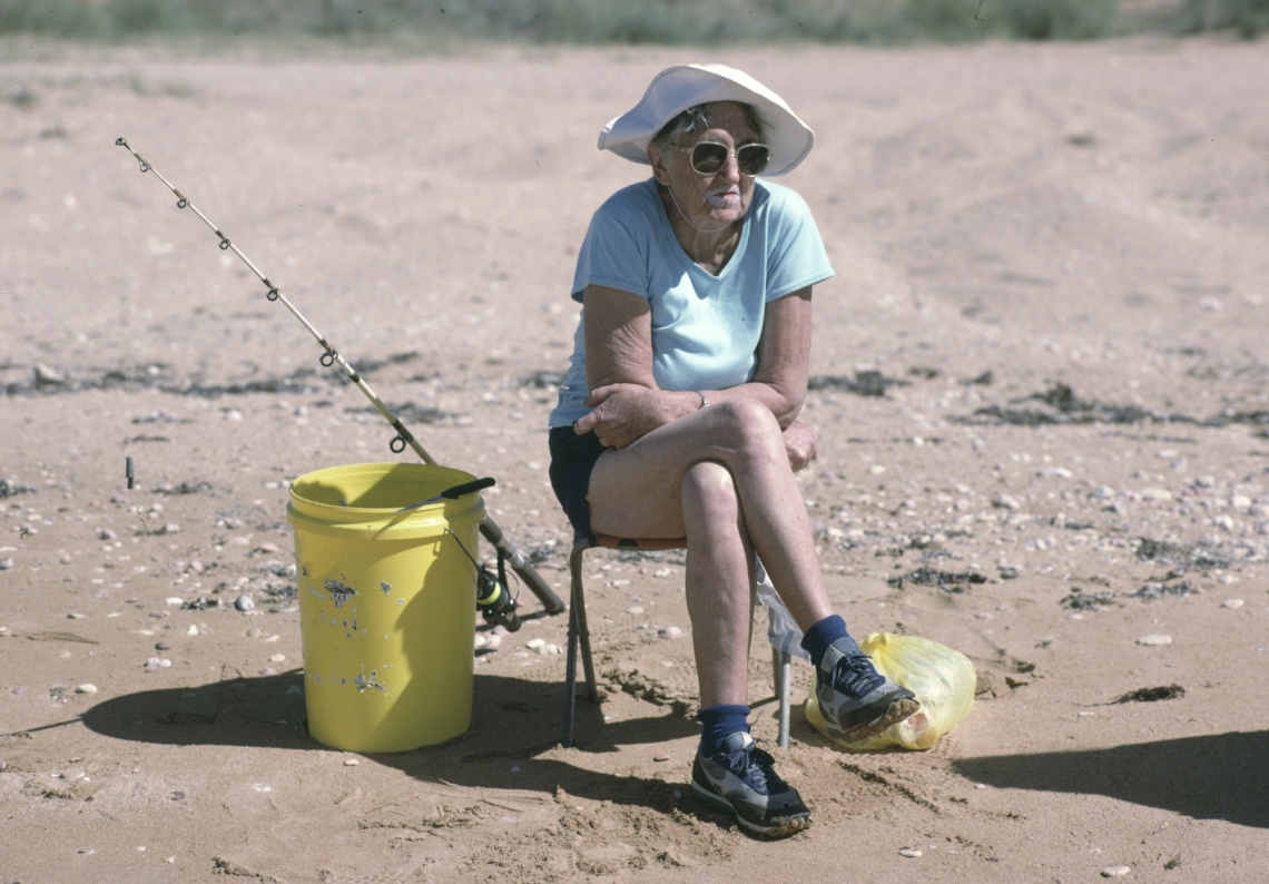 A woman sits on a stool on a beach arms crossed yellow bucket and fishing rod beside her