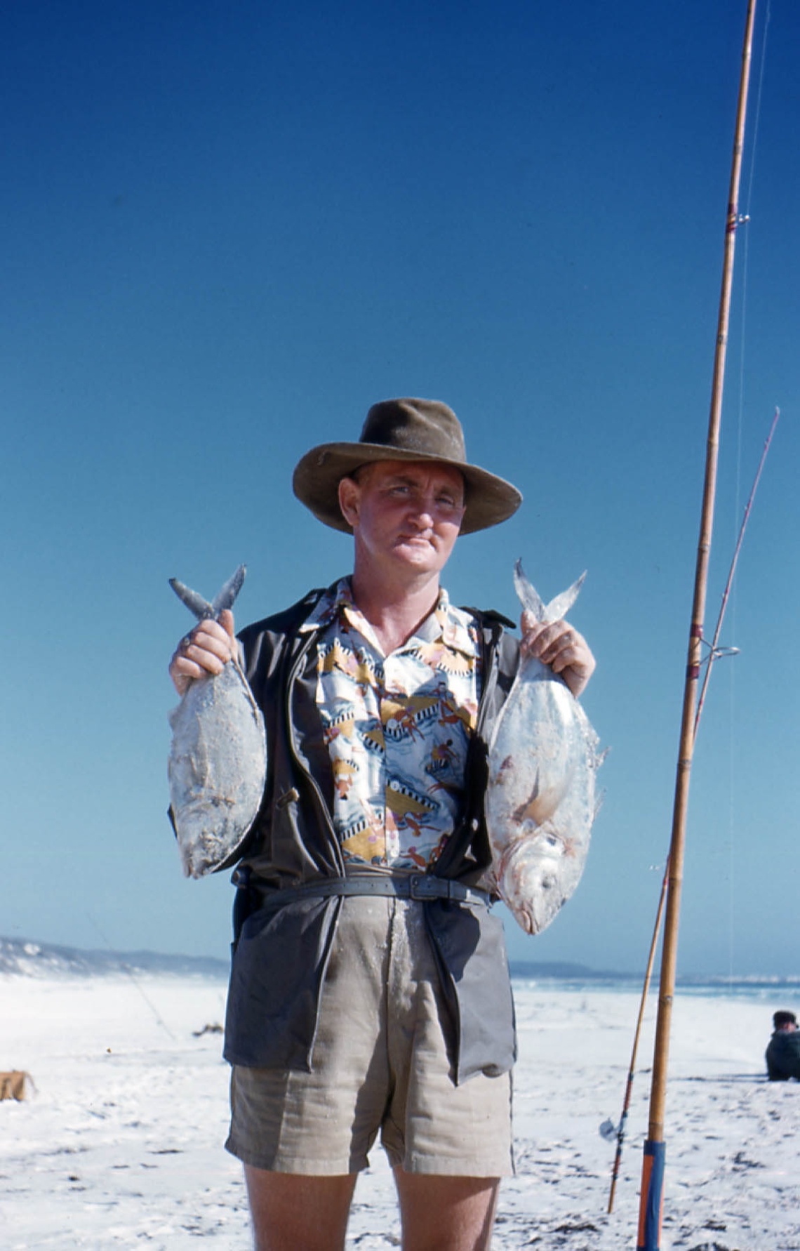 A man in a hat and shorts stands on white sand holding two silver fish by their tails