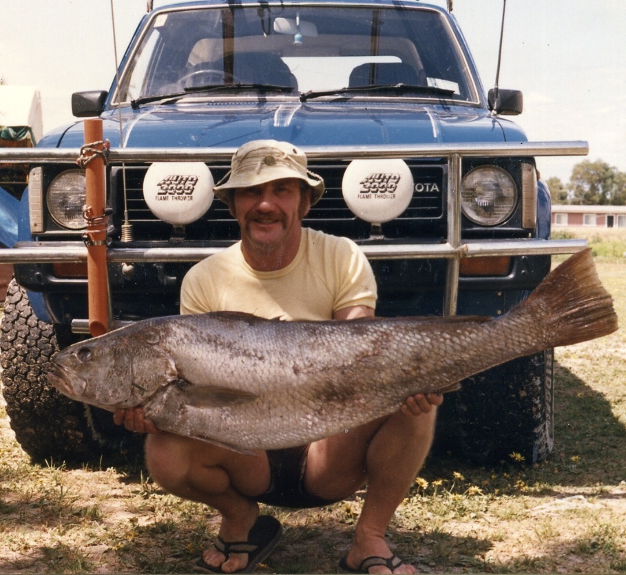 A man crouches in front of a 4wd holding a large fish almost as long as the car is wide